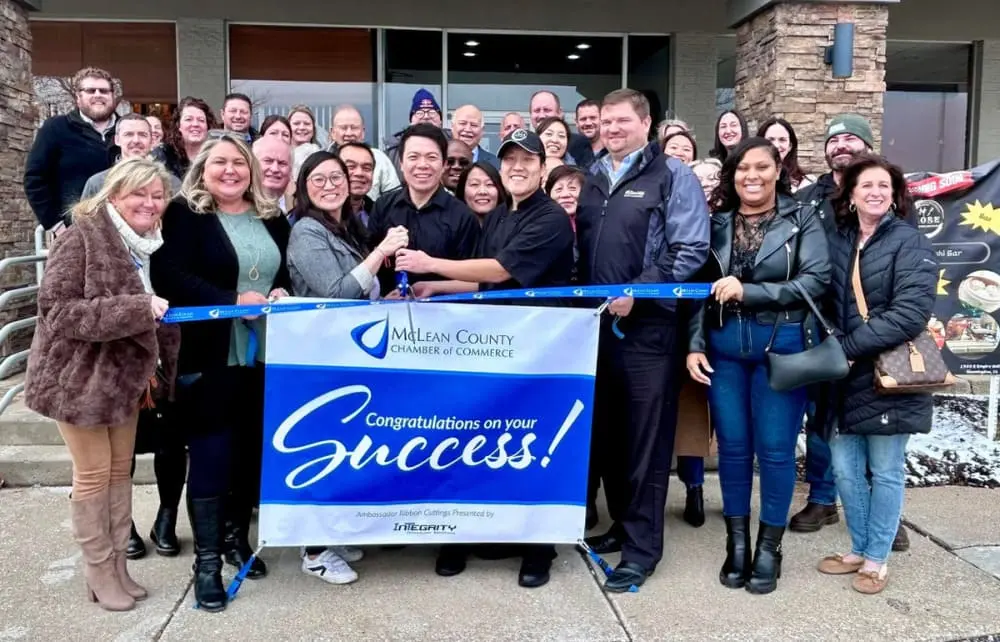 麦克林县商会成员手持红绸带举行剪彩仪式，蓝白横幅印有"Mclean County CHAMBER of COMMERCE"及"Congratulations on your Success!"字样。Integrity Bank与Bank of America标识清晰可见，体现商业社区协作。众人笑容洋溢，背景为浅色建筑。