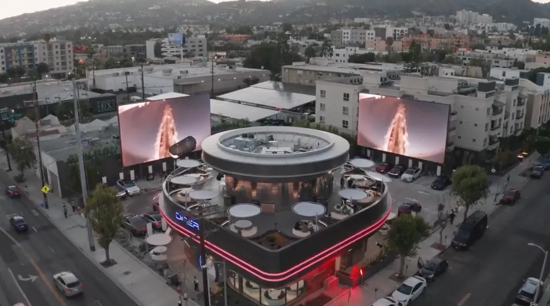 特斯拉餐厅鸟瞰全景：米白建筑顶部圆形Skypad观影台，外墙环绕红色LED灯带。两侧66英尺巨屏播SpaceX火箭影像，80个V4超充桩分布广场——全球最大城市充电站实景，背景好莱坞山强化地标属性。