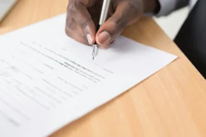 深肤手持银色钢笔签署文件，白纸聚焦"Signature"及"Financial Controller"关键字段，蓝色墨水痕迹清晰。浅灰衬衫袖口与深色外套象征商务正式性，呼应文中"退伙协议需书面化"的法律意识。