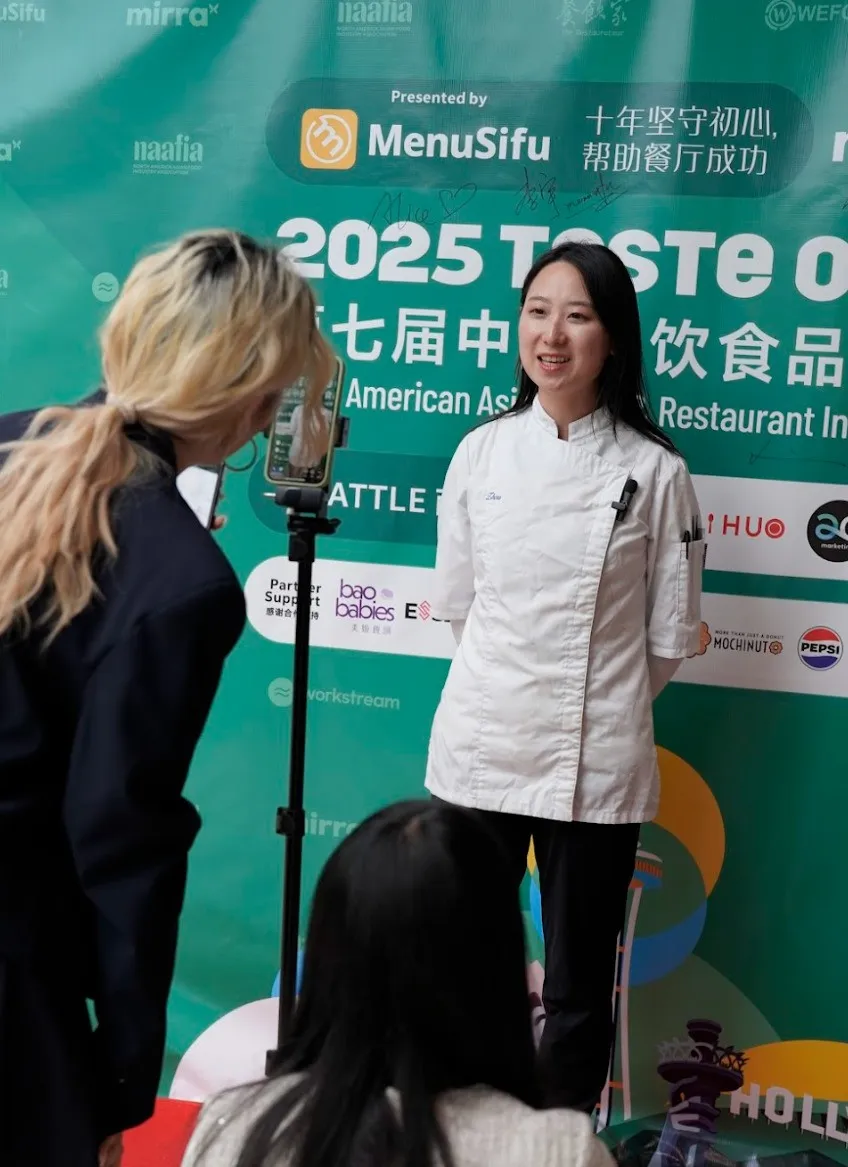 周净媛在"2025 TASTE OF ASIA"绿色展板前受访，背景含MenuSifu/海尔/大金龙等品牌标志。左侧工作人员持手机拍摄，右侧观众背影模糊虚化。活动场景印证餐饮金像奖行业影响力。
