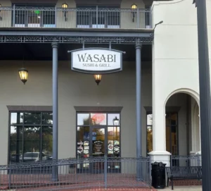 Wasabi寿司烧烤餐厅米白双层外观：① ​​黑色招牌“WASABI”​​ 配深灰立柱（象征重生起点）；② 玻璃门贴​​红色“OPEN”标​​+菜品图（疫期坚守印证）；③ 二层铁艺栏杆/复古壁灯（品质升级细节）。红砖地面呼应文中“稳扎稳打”经营哲学。