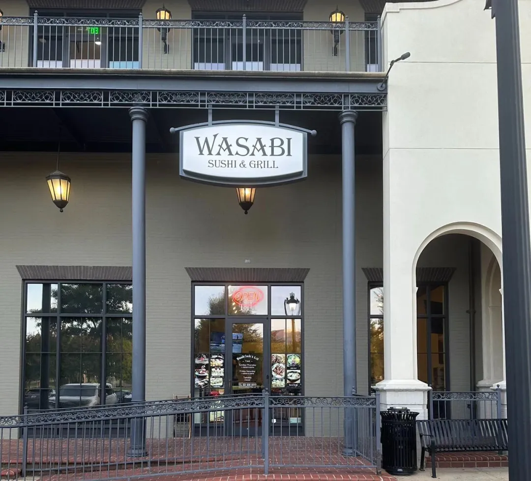 Wasabi寿司烧烤餐厅米白双层外观：① ​​黑色招牌“WASABI”​​ 配深灰立柱（象征重生起点）；② 玻璃门贴​​红色“OPEN”标​​+菜品图（疫期坚守印证）；③ 二层铁艺栏杆/复古壁灯（品质升级细节）。红砖地面呼应文中“稳扎稳打”经营哲学。