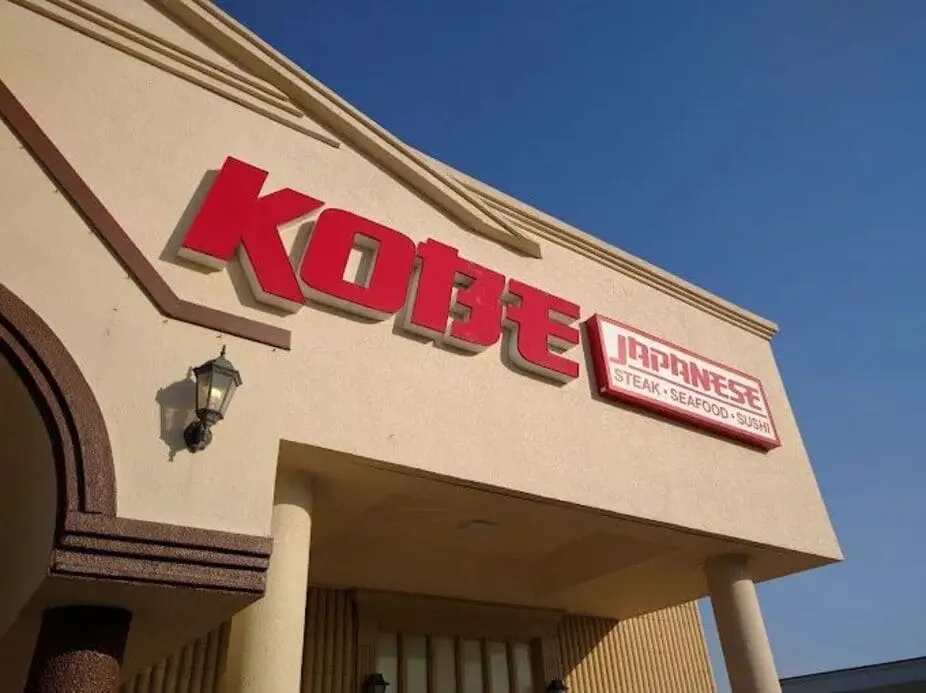 KOBE日式餐厅蓝色立体招牌特写，白色字母展示"JAPANESE STEAK·SEAFOOD·SUSHI"。米色外墙配拱形门洞与复古壁灯，建筑线条简洁，蓝天衬托日式风格。门店临街视角突显专业餐饮形象。