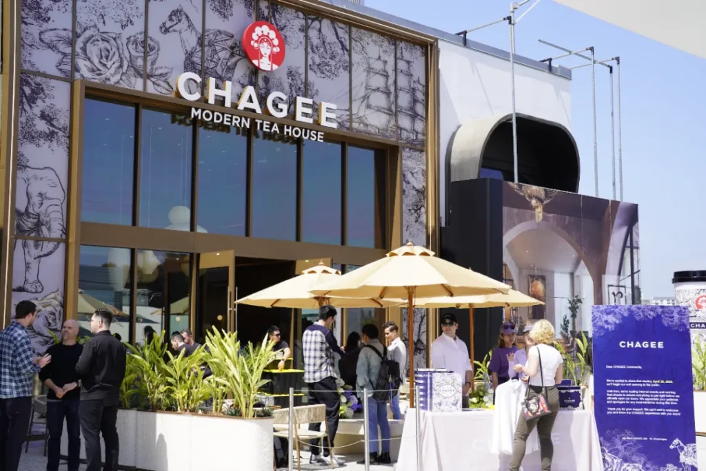 CHAGEE门店全景：大面积落地玻璃窗饰玫瑰/骏马金边艺术贴膜，顶部白底招牌嵌红唇头饰Logo。前景米白遮阳伞下顾客持饮品社交，玻璃反光映蓝天与"MODERN TEA HOUSE"标识，金色窗框构建轻奢视觉锤。印证「现代茶饮第三空间」定位。