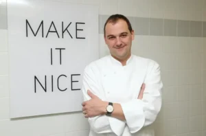 胡姆自信立于"MAKE IT NICE"标语前，瓷砖墙与黑色字体呼应EMP极简美学。此理念贯穿纯素实验至转型决策，体现主厨"创新须与现实平衡"的新哲学。