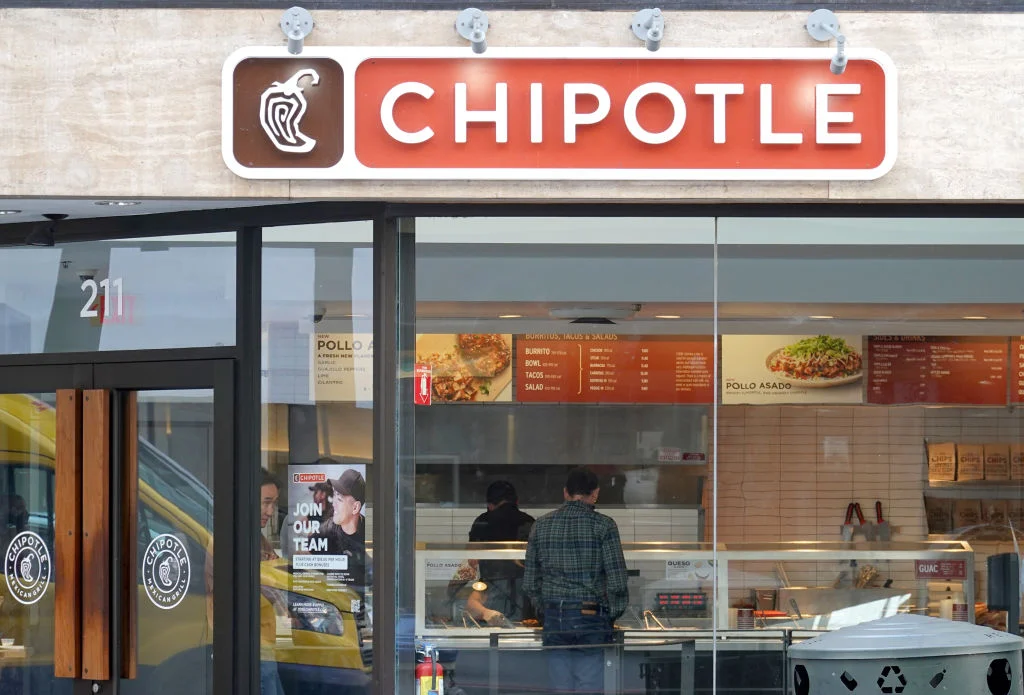 Chipotle米黄色石墙店面，橙红招牌显“CHIPOTLE”及辣椒图案；玻璃橱窗内两位顾客点餐，透过玻璃可见店内橙红菜单板（列“POLLO ASADO”等餐品）、点餐柜台及“JOIN OUR TEAM”招聘海报，光线明亮。