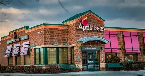 画面展示Applebee's餐厅外观，砖红色外墙配红苹果标志及白色店名，门窗有粉色遮阳篷，门前设长椅；天空暗沉营造静谧氛围，门头标语“Welcome Back!”清晰可见，凸显品牌实体形象与回归感。
