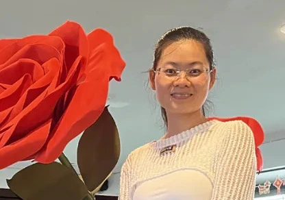 Gloria Ye戴细框镜微笑倚靠巨型红玫瑰茎杆，白针织衫显知性气场。花茎土壤层埋入云南普洱茶饼与草果香料，根系蔓延象征心性根基，印证"供应链精益源于信念"论述。深棕背景强化沉浸氛围，呼应"稳定灵魂成就品牌一致性"观点。
