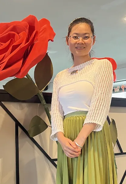 Gloria着绿裙立于绽放巨玫瑰前,双手交叠显从容仪态。花瓣飘落轨迹化为"70%"复购率数据流,融入顾客笑脸虚影,具象化"频率吸引精准客群"飞轮模型。暖光穿透花瓣纹理,暗喻"厚德载福善业聚财"能量磁场辐射社区。