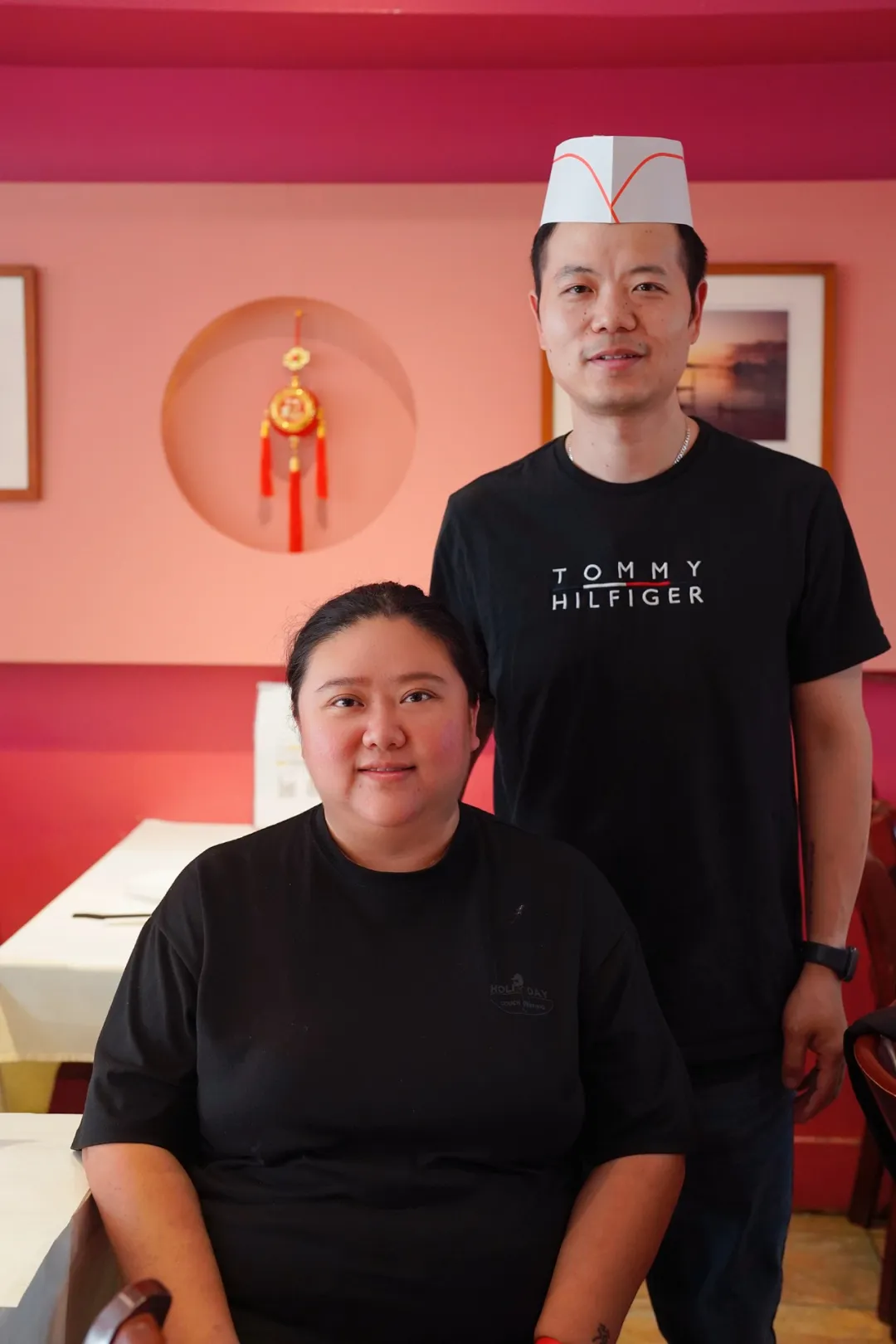 男女在粉墙中式装饰餐厅内合影，女坐黑衣，男戴白帽穿Tommy HilfigerT恤，氛围温馨亲切。
