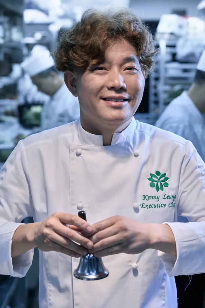 金像奖杰出厨师得主梁继坚主厨特写：身穿绣"Kenny Leung Executive Chef"白色制服，手托餐饮铃笑容自信。胸前三叶草徽章象征"清、新、鲜"烹饪哲学，背景厨房团队忙碌场景虚化处理，突出人物权威性。