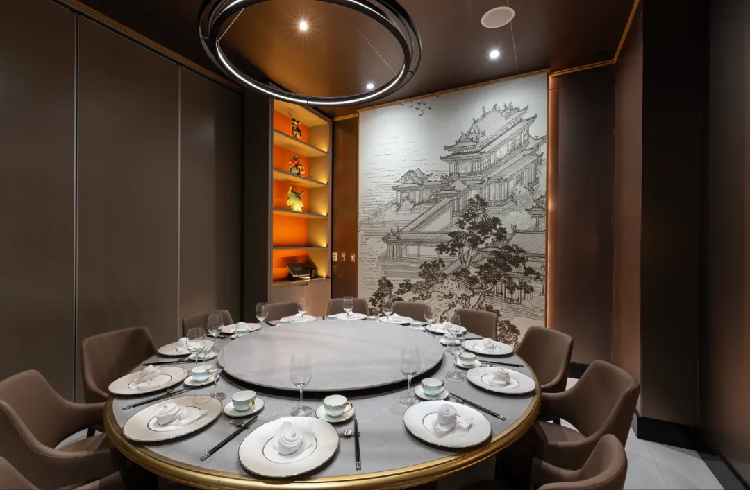 富瑶华尔街店中式包厢全景：圆形餐桌铺白色餐布，配金色镶边骨瓷餐具。背景墙手绘宋代风格建筑群壁画，左侧仿古置物架悬暖橙射灯，顶部环形水晶吊灯映射传统与现代交融理念。座椅采用深咖皮革材质，营造商务宴请氛围。