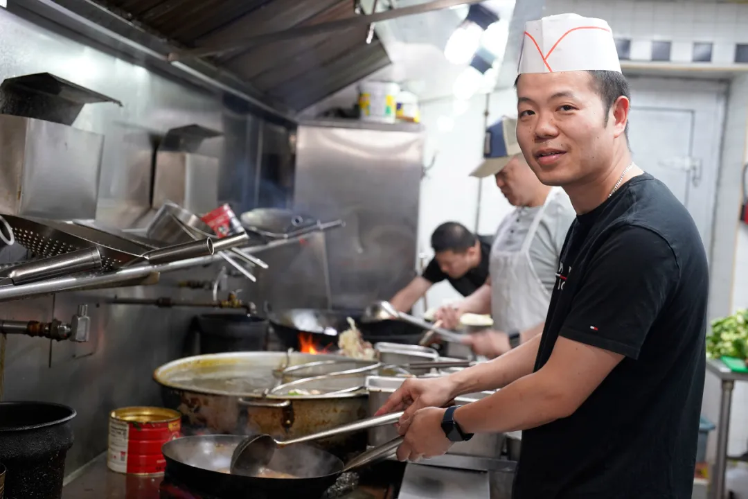戴白帽黑衣男子在厨房操作台前持夹具备餐，身后其他工作人员忙碌烹饪，展现后厨烟火气。