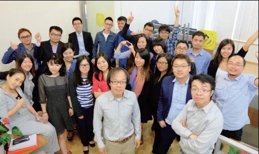 约25人团队在明亮办公室合影，前景男士穿浅色衬衫，后排人群或站或坐，部分做胜利手势，背景有玻璃隔断、黄色标志，氛围轻松愉快。