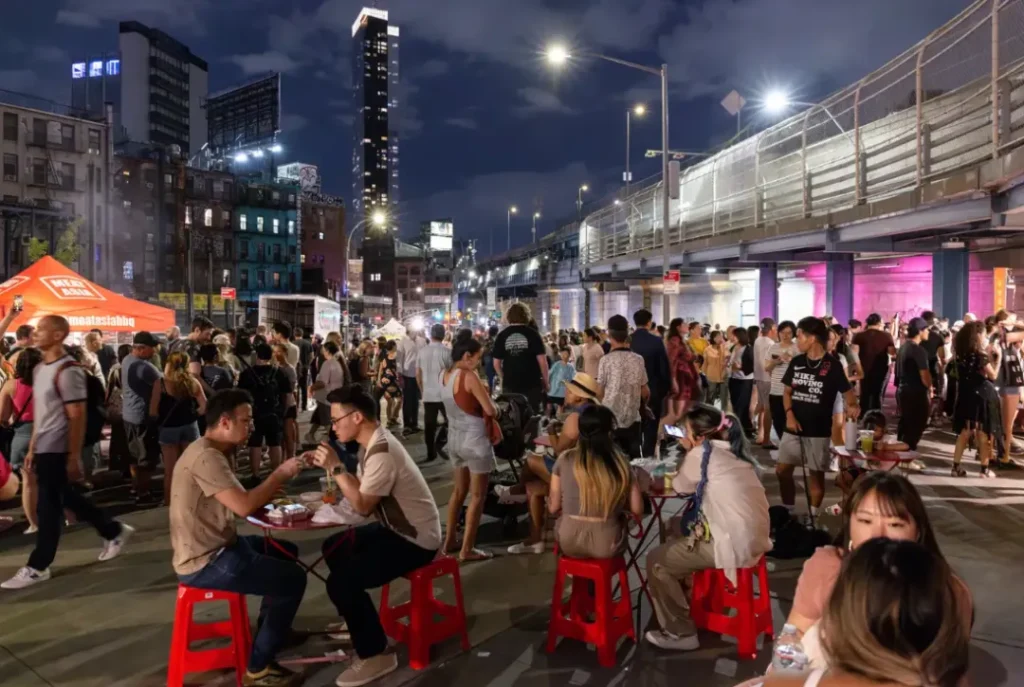 曼哈顿大桥橙光笼罩夜市：左侧帐篷摊位悬挂“Asia Food”灯牌（规避水印文字），售卖中式包子/尼泊尔momo饺子。前景木桌人群举烤串碰杯，后方高楼林立呼应文中“都会夜生活场域”。红灯笼串点缀夜空，取证Think!Chinatown通过多元美食重建社区安全感，关联2025年8月终场活动。
