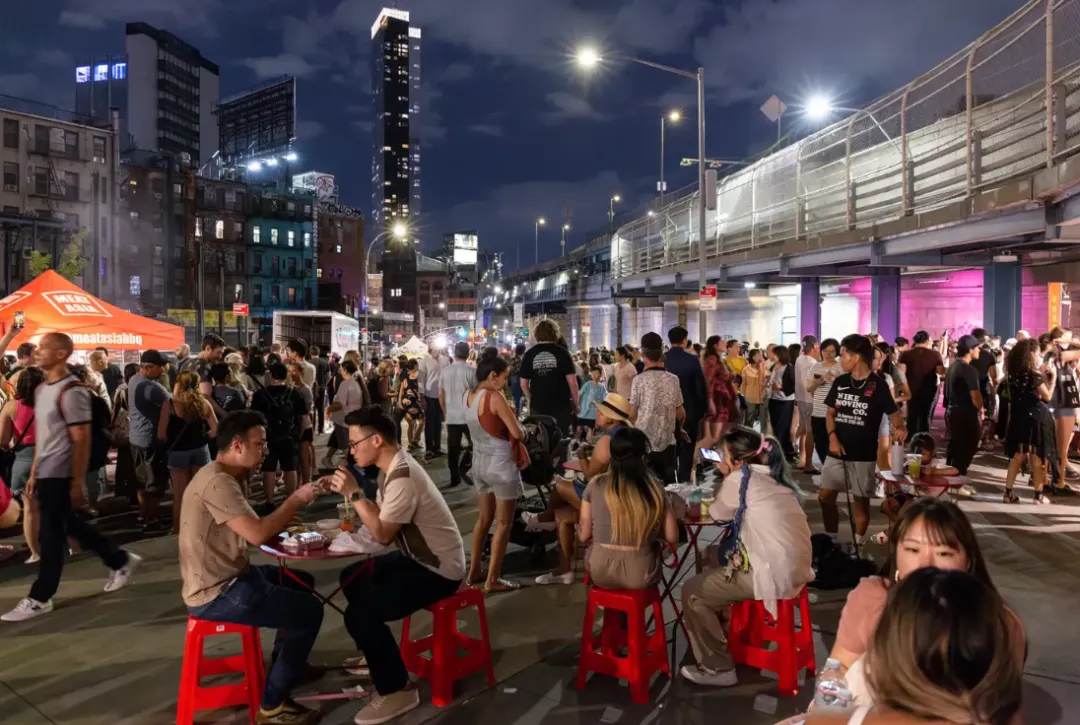 曼哈顿大桥橙光笼罩夜市：左侧帐篷摊位悬挂“Asia Food”灯牌（规避水印文字），售卖中式包子/尼泊尔momo饺子。前景木桌人群举烤串碰杯，后方高楼林立呼应文中“都会夜生活场域”。红灯笼串点缀夜空，取证Think!Chinatown通过多元美食重建社区安全感，关联2025年8月终场活动。