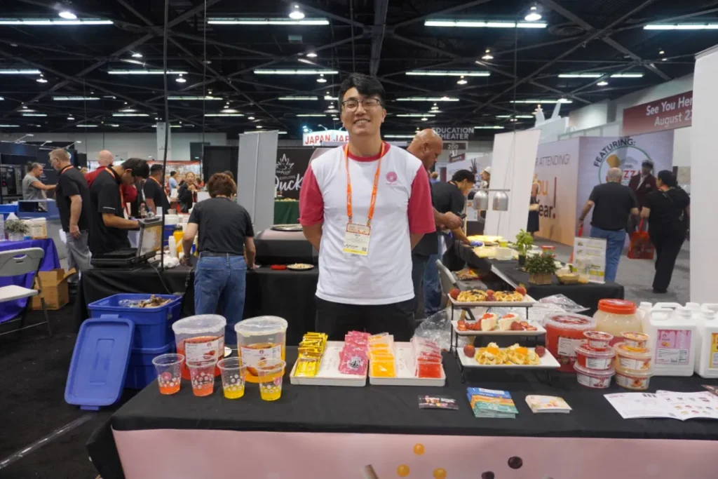 展位工作人员（戴眼镜穿红白T恤）讲解设备功能，背景可见商用披萨烤箱（HEATER 200型号）、日本进口烹饪设备（JAPAN JAP标识）及饮品展示台。左侧设备屏显"FEATURING ATTENDING"，印证餐饮设备智能化趋势，呼应文中"RoboOp365机器人"技术展示内容。