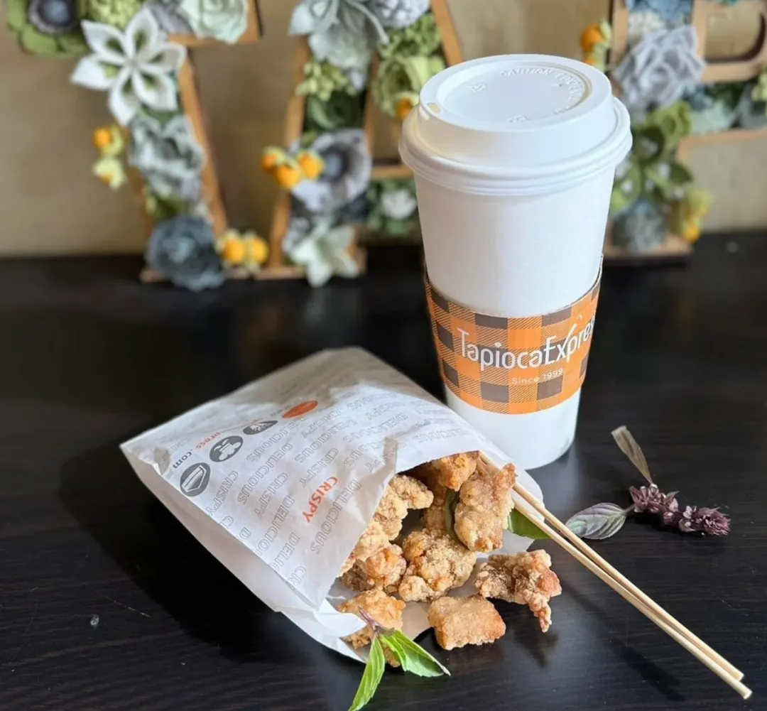 Tapioca Express品牌特写：前景为金黄酥脆的盐酥鸡块散落在白色印花纸袋上，搭配带橙色格纹杯套的珍珠奶茶。木签穿插鸡块间，新鲜罗勒叶点缀桌面。虚化背景可见"T""E"木质字母与花艺装饰，强调"CRISPY DELICIOUS"质感与1999年创立品牌历史。