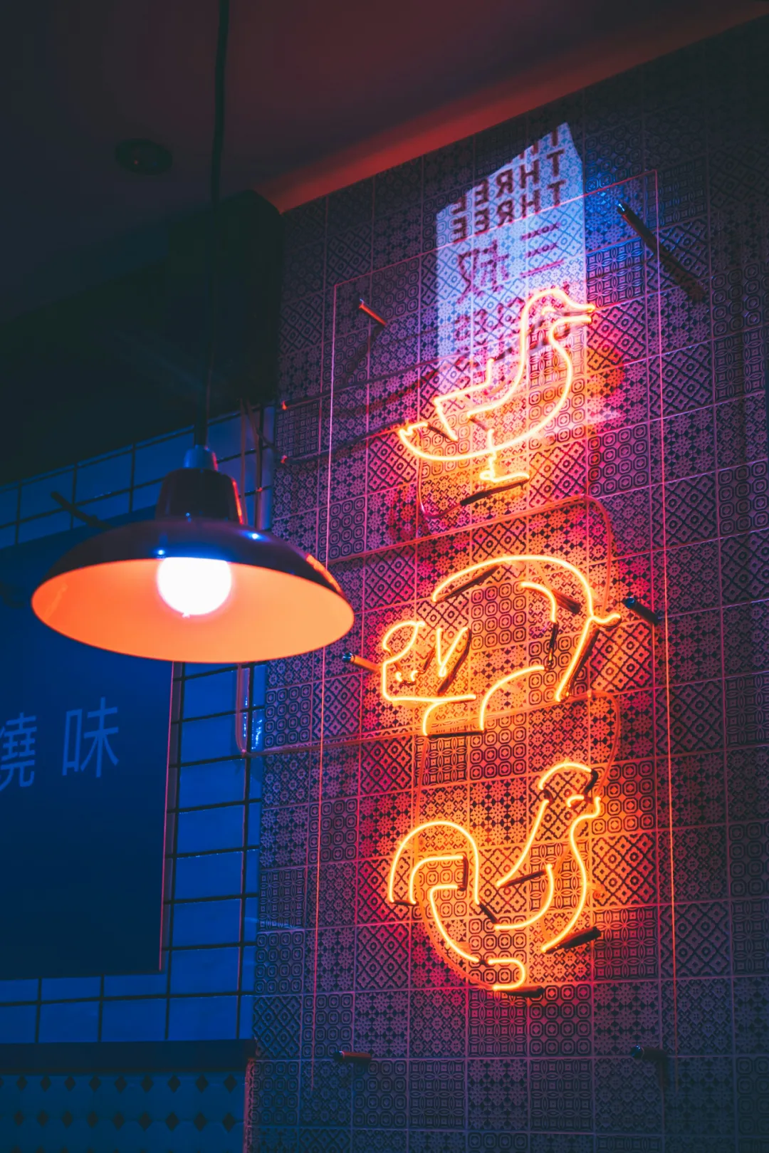 烧腊店装饰墙特写：右侧红黄霓虹管勾勒鹤形轮廓（规避敏感文字），右下暖光吊灯在花纹瓷砖投放射状光斑。顶部倒映"THREE"冷光字样，左侧墙面肌理如焦糖脆皮。印证文中「品牌视觉锤降低获客成本」的加盟视觉优势，橙色主调呼应功夫茶案例。