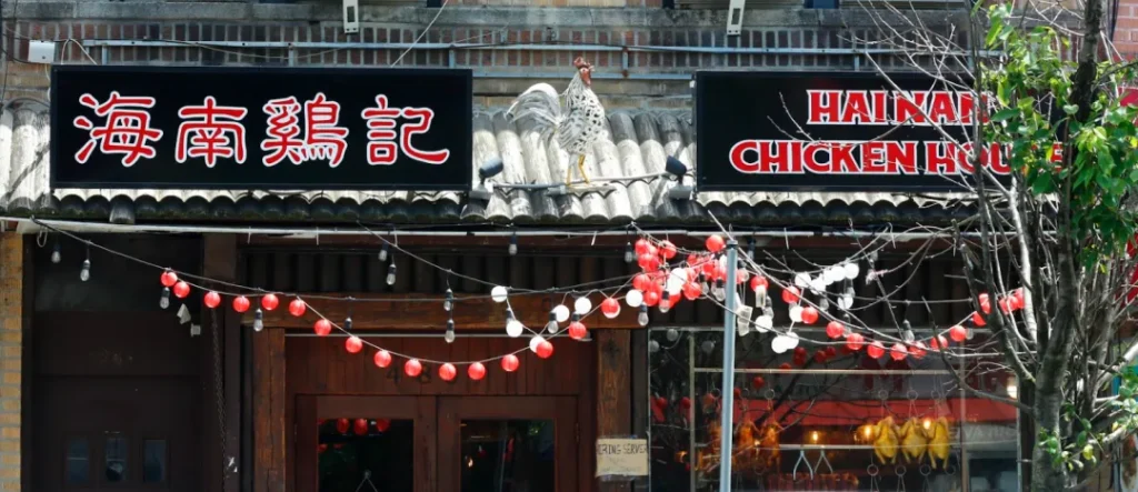黑底金字"海南鸡记"招牌（中英双语），门楣白公鸡雕塑振翅欲飞。檐下红白灯笼与暖黄灯泡串构成几何光影，棕木门透出厨房暖光。右侧橱窗烤鸡剪影呼应彩釉马来瓷砖外墙，诠释导演Chris"极简空间承载文化记忆"的设计哲学。