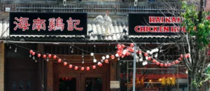 黑底金字"海南鸡记"招牌（中英双语），门楣白公鸡雕塑振翅欲飞。檐下红白灯笼与暖黄灯泡串构成几何光影，棕木门透出厨房暖光。右侧橱窗烤鸡剪影呼应彩釉马来瓷砖外墙，诠释导演Chris"极简空间承载文化记忆"的设计哲学。