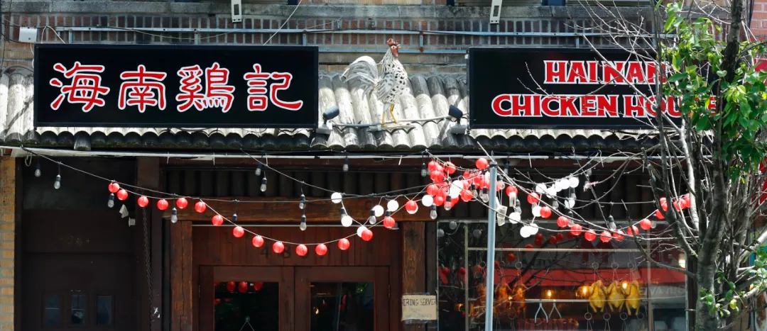 黑底金字"海南鸡记"招牌（中英双语），门楣白公鸡雕塑振翅欲飞。檐下红白灯笼与暖黄灯泡串构成几何光影，棕木门透出厨房暖光。右侧橱窗烤鸡剪影呼应彩釉马来瓷砖外墙，诠释导演Chris"极简空间承载文化记忆"的设计哲学。