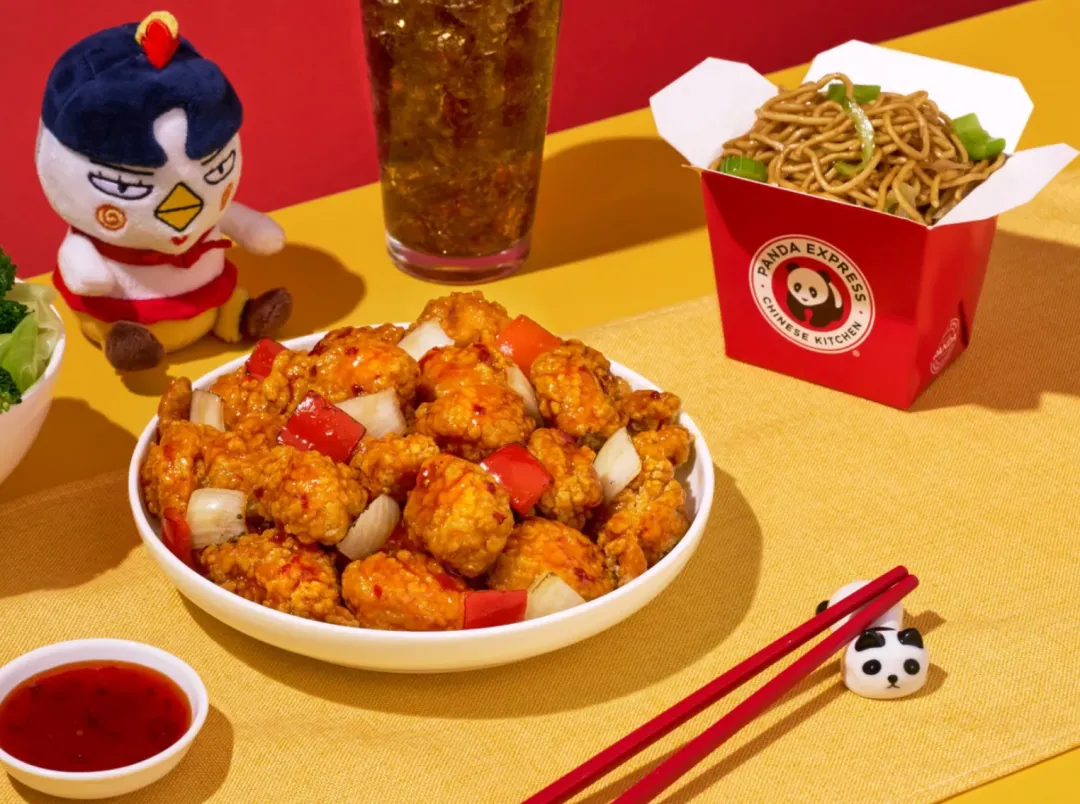 熊猫快餐（PANDA EXPRESS）标志性红黄餐盒组合：前景为琥珀色宫保鸡丁配红椒青葱，佐秘制辣椒蘸酱；右侧红色餐盒盛装豉油炒面，背景含品牌经典波霸饮品及吉祥物熊猫玩偶。炸鸡金黄酥脆、面条油润诱人，突出"CHINESE KITCHEN"美式中餐特色。