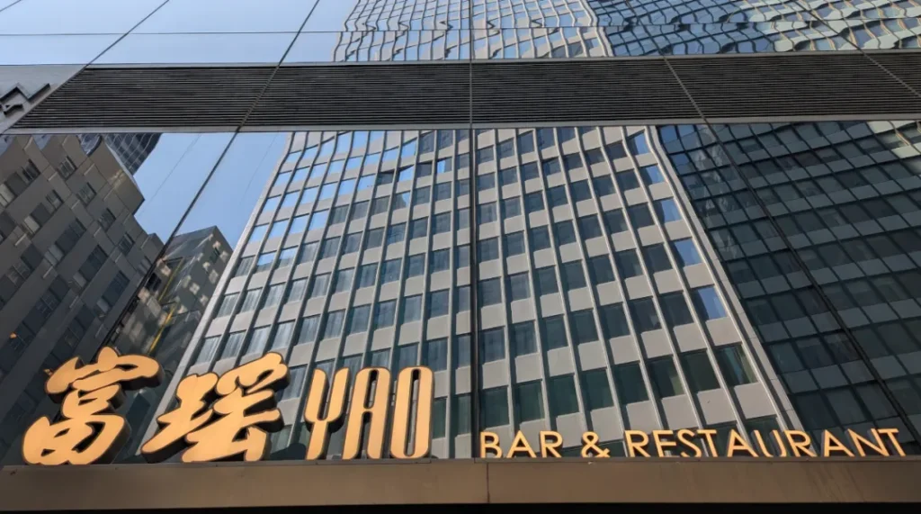 富瑶华尔街旗舰店"YAO BAR & RESTAURANT"金色立体字门头，安装于万豪酒店大理石外立面。背景玻璃幕墙反射金融区摩天楼群，下方"BAR"字样点明酒吧功能，印证"高端粤菜进驻金融核心区"战略。
