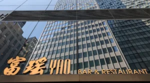 富瑶华尔街旗舰店"YAO BAR & RESTAURANT"金色立体字门头，安装于万豪酒店大理石外立面。背景玻璃幕墙反射金融区摩天楼群，下方"BAR"字样点明酒吧功能，印证"高端粤菜进驻金融核心区"战略。