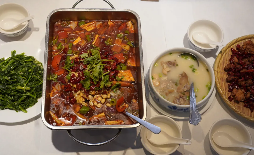 俯拍长桌丰盛川菜，中心红油麻辣香锅撒香菜花生，旁有清炒菠菜、蹄花汤及干煸肉菜，色彩诱人。