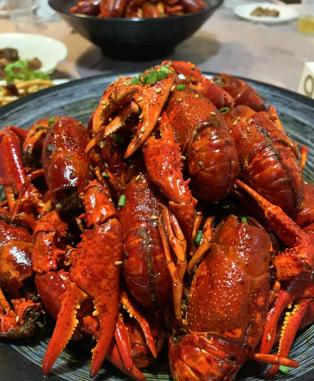 近摄深色盘中美味小龙虾，红亮油润外壳缀芝麻葱花，虾钳微张，背景模糊可见其他餐食，视觉诱人。