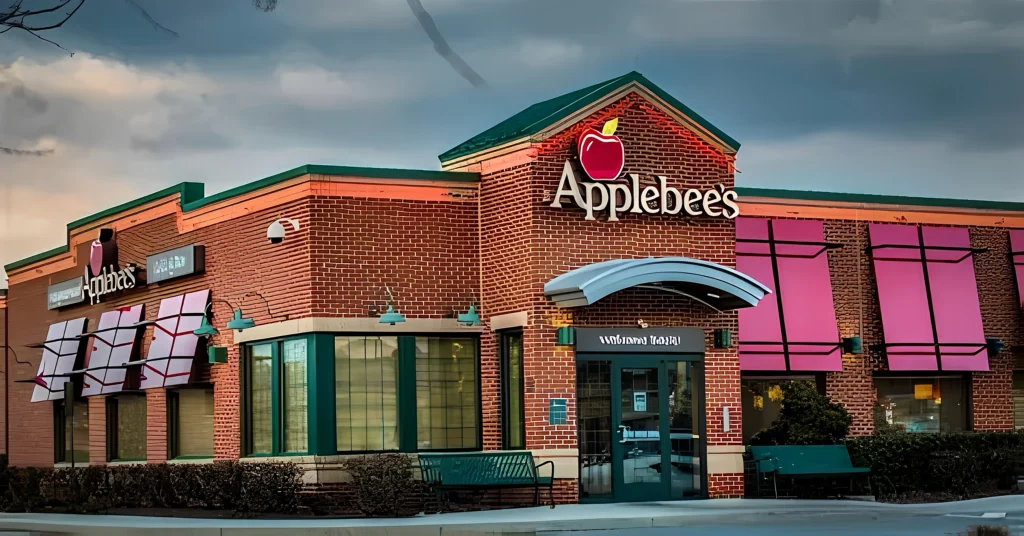 画面展示Applebee's餐厅外观，砖红色外墙配红苹果标志及白色店名，门窗有粉色遮阳篷，门前设长椅；天空暗沉营造静谧氛围，门头标语“Welcome Back!”清晰可见，凸显品牌实体形象与回归感。