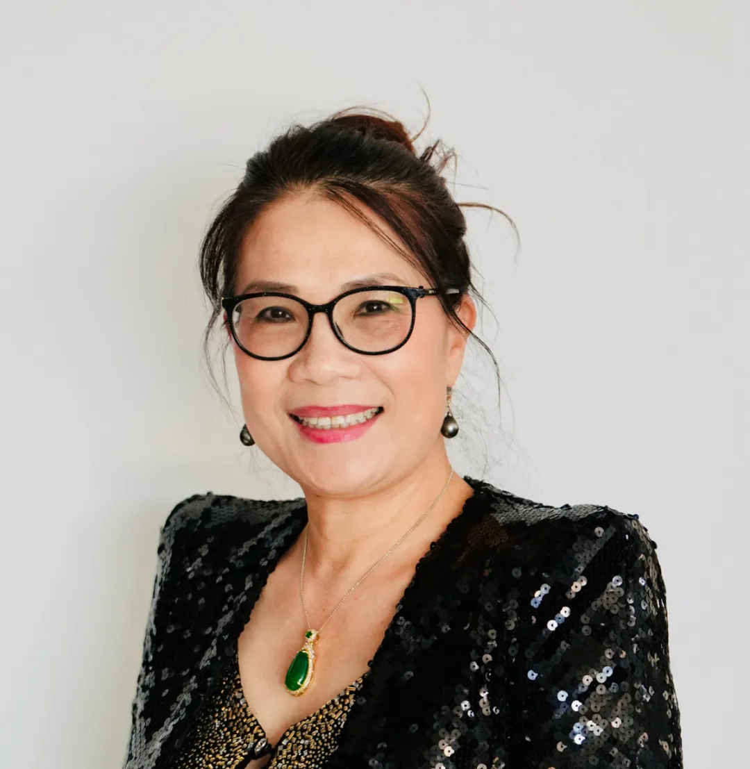 女士扎发戴黑框眼镜，穿黑色亮片外套，内搭金色花纹服饰；脖颈挂翡翠项链，耳饰黑色，面带微笑，背景简洁白色，亲切自然。