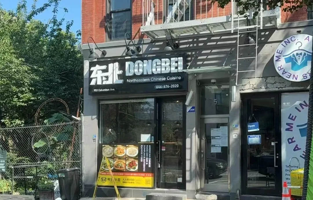 哥伦布大道“东北 DONGBEI”餐厅门面，招牌含中英文店名、地址及电话，邻裁缝店，绿植围栏点缀。