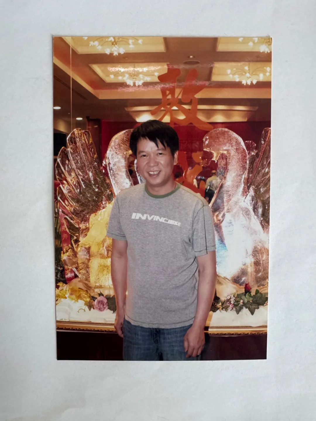 男子穿灰短袖、蓝牛仔裤，站装饰华丽场所前微笑。背景有发光天鹅装饰、红色汉字与璀璨灯光，氛围喜庆。