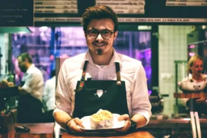 戴眼镜男性服务员穿白衬衫黑围裙，双手端食物盘微笑看向镜头，背景为忙碌餐厅内景，灯光温馨，展现服务温馨氛围。