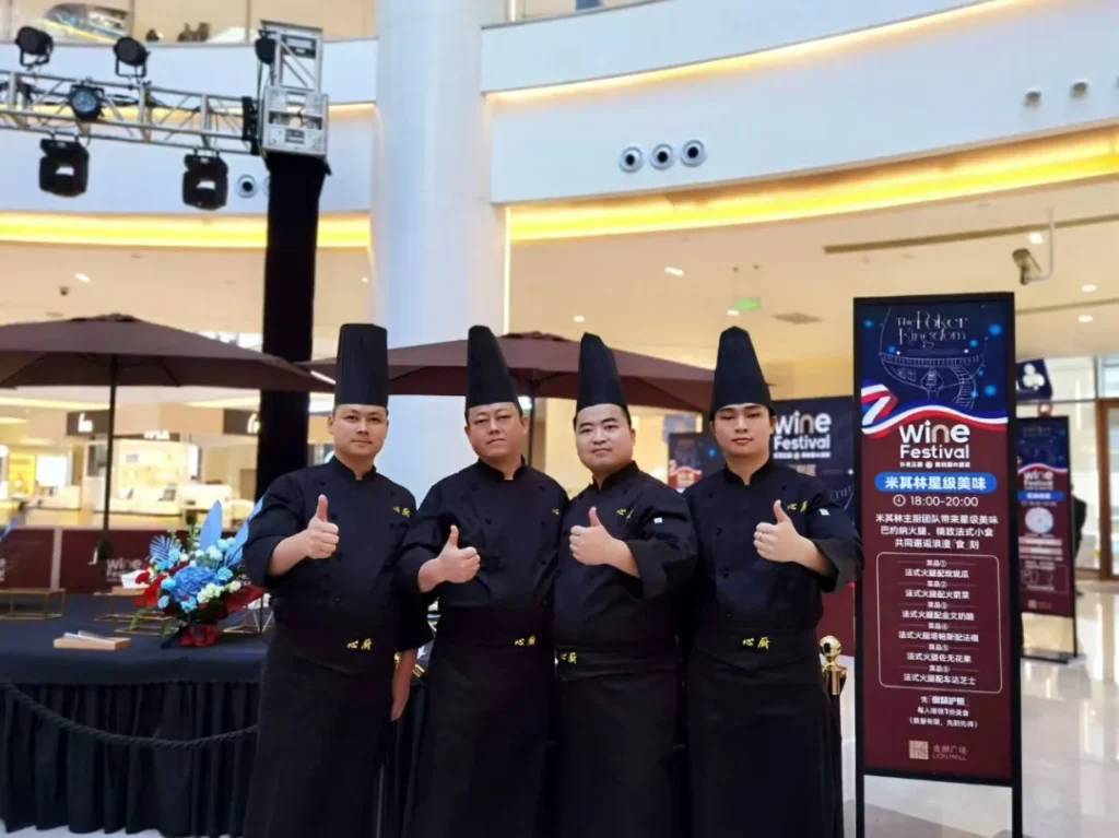 四位穿黑色厨师服戴高帽者站一排竖大拇指，身后有“Wine Festival 20周年”展板、桌椅鲜花，氛围热闹活动现场。