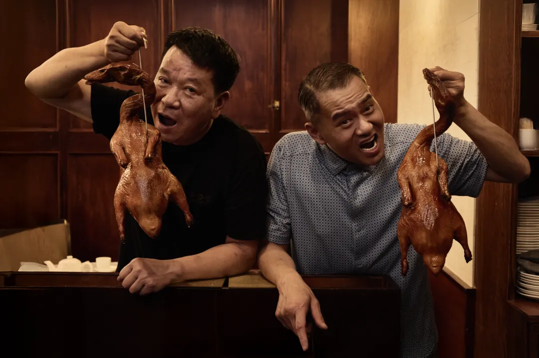 两位男子在木质装潢餐馆内，分别举着下垂的烤鸭，表情兴奋。左穿黑短袖，右穿蓝底白点短袖，靠深色台面，氛围热闹生活化。