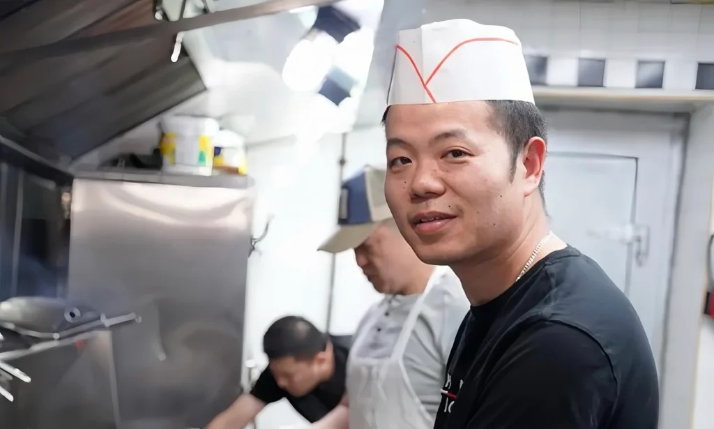 戴白帽黑衣男子在厨房操作台前持夹具备餐，身后其他工作人员忙碌烹饪，展现后厨烟火气。