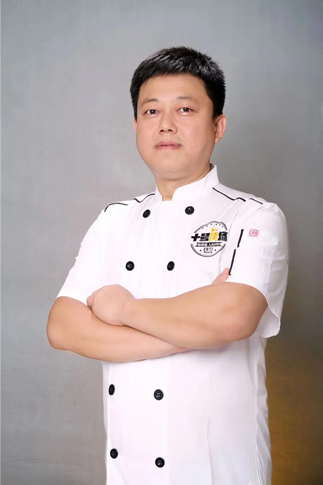 穿白色厨师制服男性双臂交叉站立，左胸印“十号烤场BBQ LAND 2011”徽章，背景灰色渐变，表情沉稳，职业感十足。