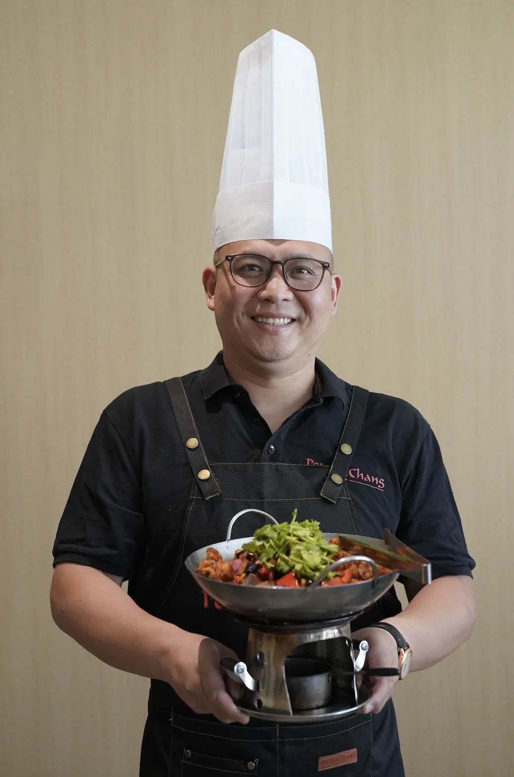 戴眼镜中年男厨师穿黑Polo衫、深色“Chans”围裙，微笑端金属干锅；锅内肉丁、红椒、黑豆豉配翠绿香菜，下有酒精炉加热，背景浅褐竖纹墙。