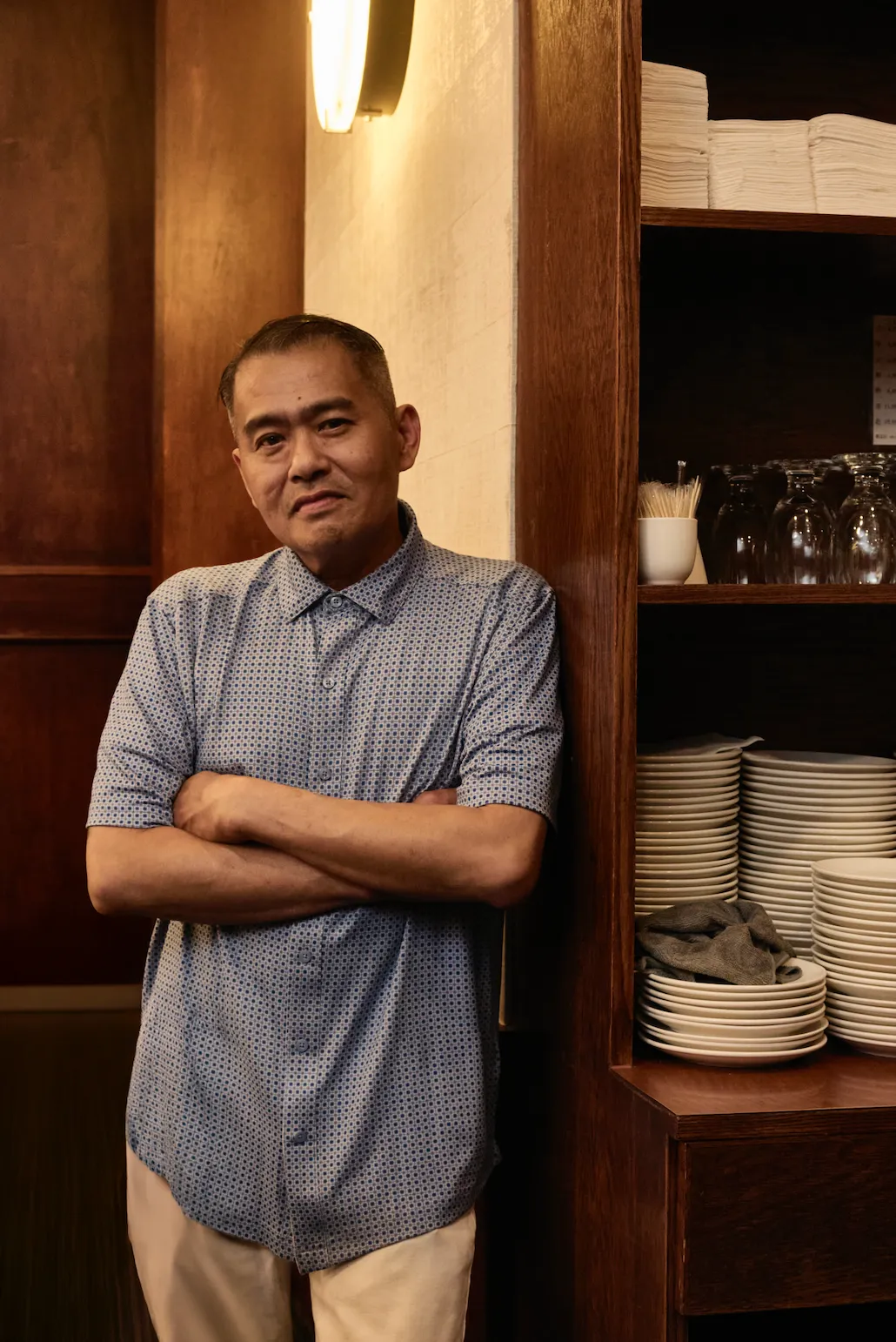 男子穿蓝底花纹衬衫、米色裤，双臂交叉站深棕木质置物架旁。架上有盘子、玻璃杯，背景暖木墙与壁灯，光线柔和显专业氛围。