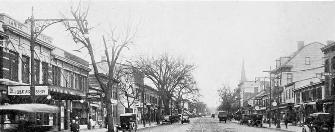 黑白照片，旧城市街道两旁多层建筑，底层商铺招牌清晰，有老式汽车、有轨电车和行人，树木点缀，呈现旧时繁忙日常。