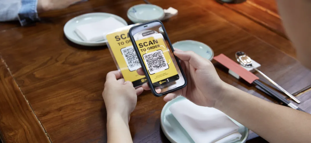 餐厅内，一双手持手机扫描餐桌上的“SCAN TO ORDER”二维码，桌上有木质餐盘、餐具与餐巾，背景隐约可见他人腿部，呈现现代便捷点餐场景。