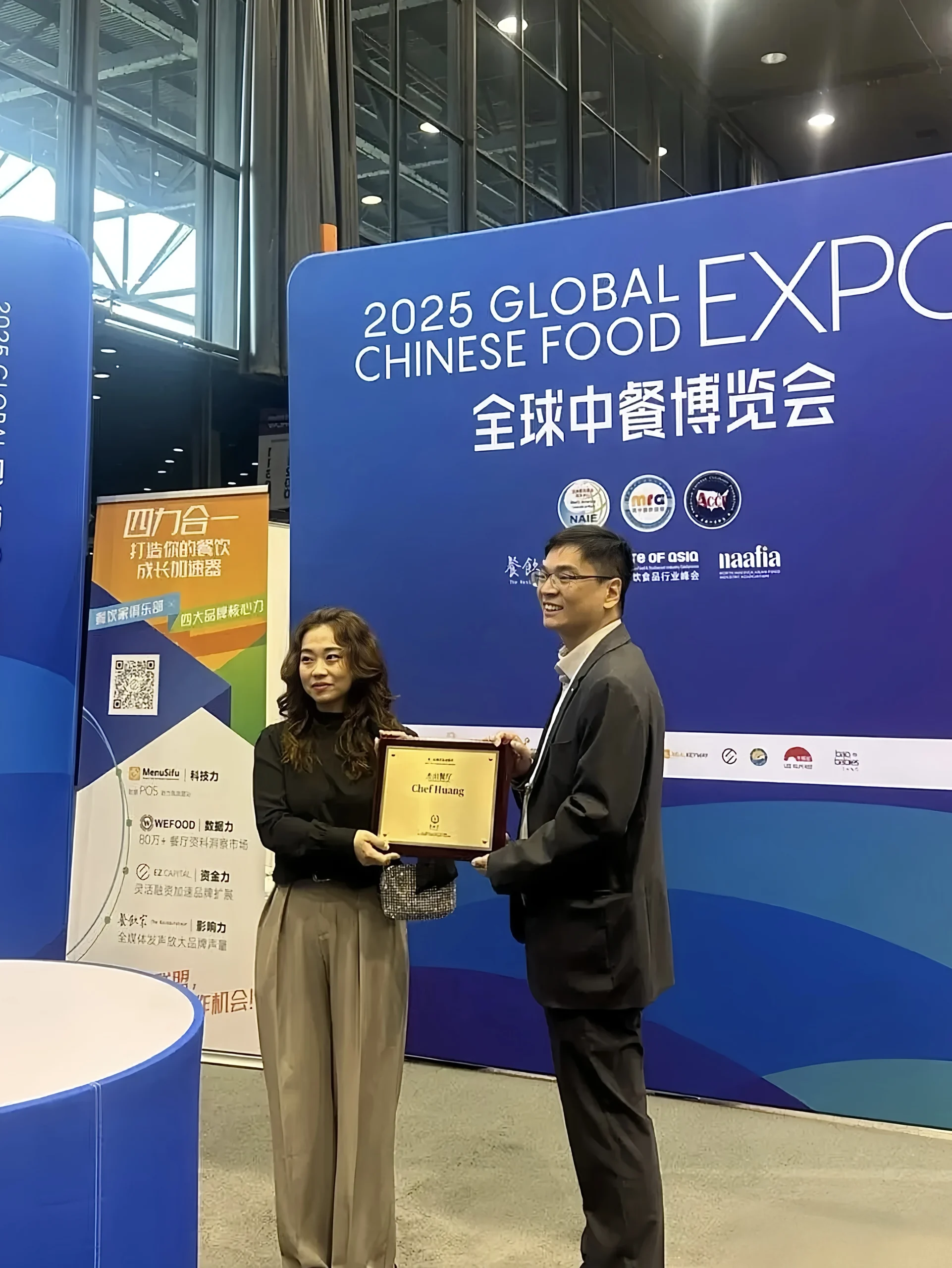 在2025全球中餐博览会现场，李锦记美东负责人 Kam Shek 正式为 Chef Huang 颁发杰出餐厅奖。两人在展会舞台前合影留念，手中展示着印有获奖者姓名的奖牌。