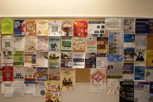 浅色墙面棕色软木板公告栏，贴满语言交流、志愿者招募、求职信息等彩色海报，设计风格多样（简洁/图文并茂），整体充满生活气息。