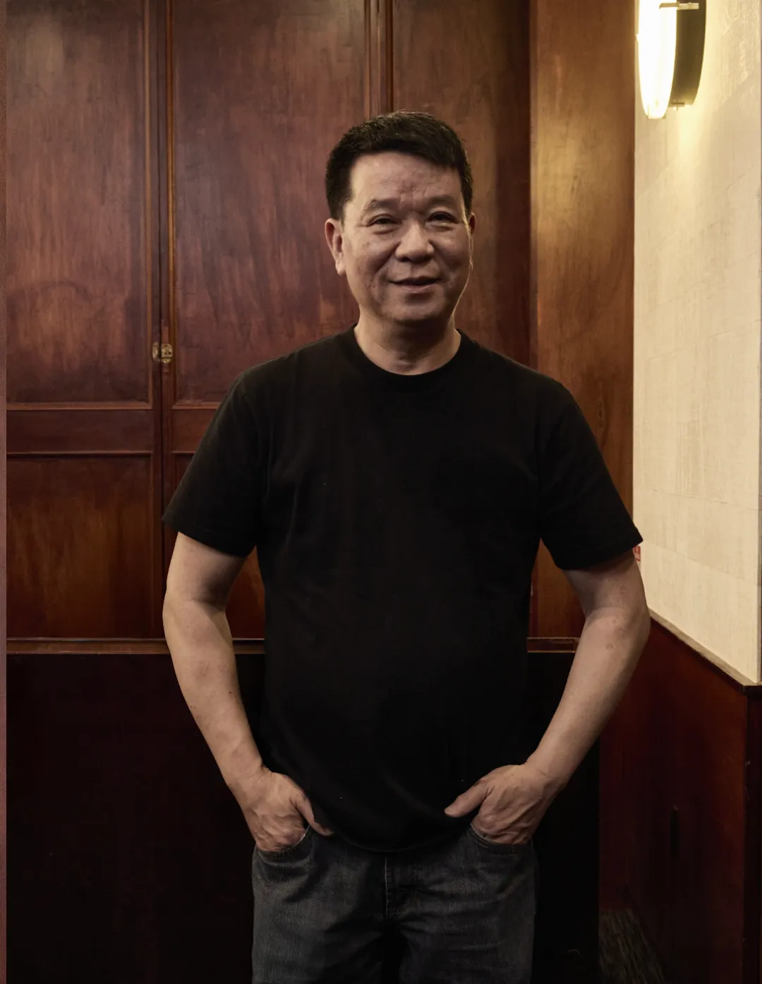 中年男子穿黑色短袖T恤，双手插牛仔裤袋，微笑看镜头。背景木质墙面配壁灯，氛围温馨亲切，姿态自然随和。