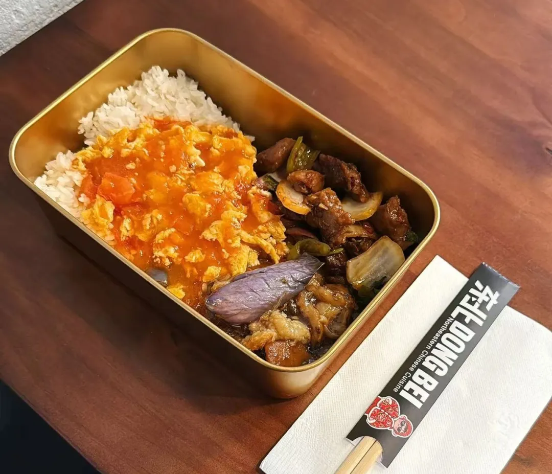 木质桌面摆金色餐盒，内盛番茄炒蛋饭与肉茄菜肴，配筷子及印“东北 DONG BEI”字样餐巾，呈现丰盛中式便当。