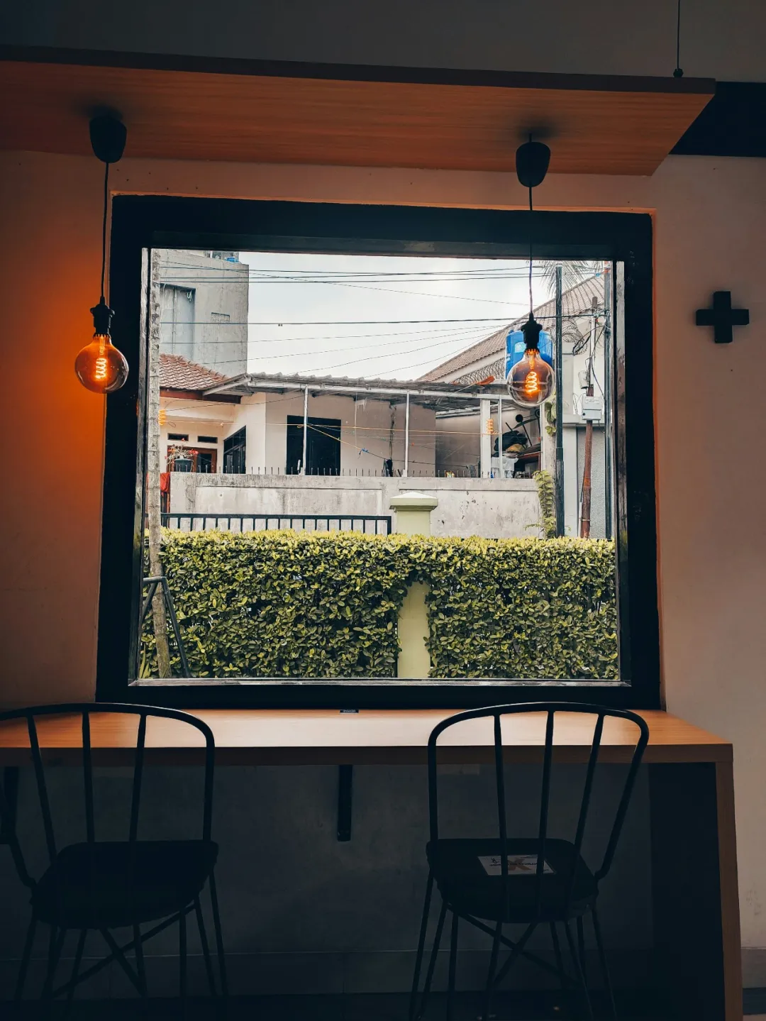 室内窗边，大窗透出户外绿色树篱与房屋，木质桌配黑色金属椅，两侧挂暖黄复古灯泡，浅色墙与十字装饰点缀，灯光营造静谧温馨氛围。
