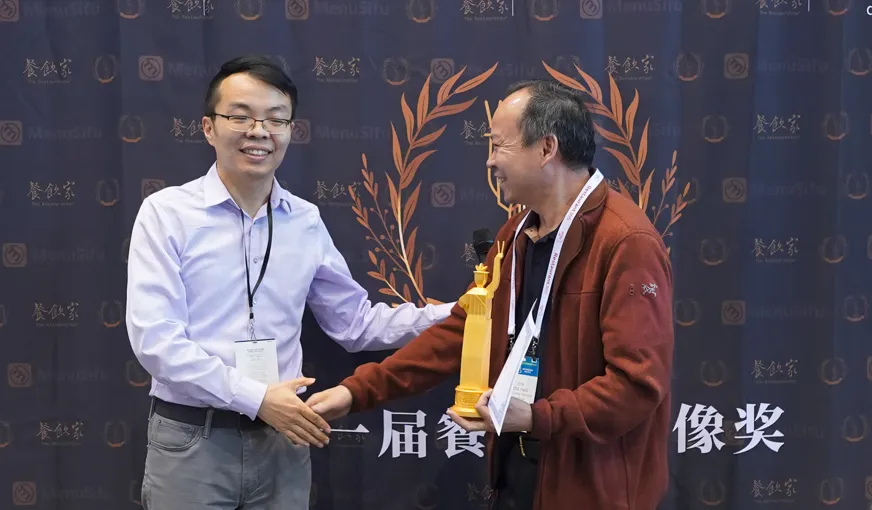 浅紫衬衫戴眼镜男士（左）微笑搭红棕外套男士（右）手臂，右男手持金色奖杯与蓝边证书，胸前挂证件，背景为深蓝“餐饮家”幕布，带金色枝叶图案。